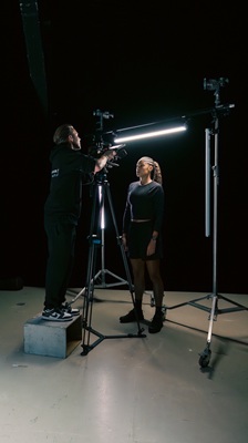 Eine Person steht auf einem Kasten und richtet eine Kamera auf die Fussballerin Sydney Schertenleib, die vor einem schwarzen Hintergrund steht. Es handelt sich dabei um eine behind the scenes-Aufnahme für eine Werbefilmproduktion für die Uhrenmarke Norqain produziert im Mietstudio für Film- und Fotoproduktionen StudioGutenswil in der Nähe von Zürich (Schweiz).