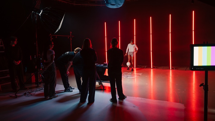 Eine Gruppe von Personen in einem Studio mit roten Lichtstreifen, während die Fussballerin Sydney Schertenleib mit einem Fussball jongliert. Es handelt sich dabei um eine behind the scenes-Aufnahme für eine Werbefilmproduktion für die Uhrenmarke Norqain produziert im Mietstudio für Film- und Fotoproduktionen StudioGutenswil in der Nähe von Zürich (Schweiz).